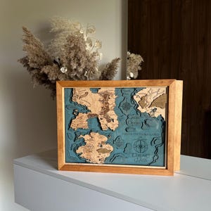 May include: A wooden map of the fictional world from the TV show Game of Thrones, with a blue ocean and brown landmasses. The map is framed in a light brown wood frame. The text "Game of Thrones" is visible in the lower right corner of the map.