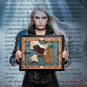 May include: A framed wooden map with intricate details, featuring landmasses in shades of brown and green against a blue background. The frame is black, and the map has decorative elements. The overall theme is fantasy or adventure.
