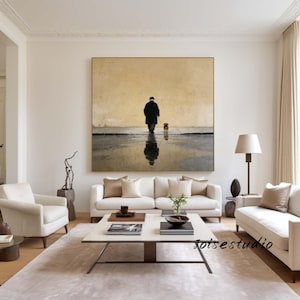 May include: A large framed painting depicts a person walking a dog on a wet surface, with the person's reflection visible. The artwork is in a neutral color palette, with a beige background and dark figures. The painting hangs above a white sofa.