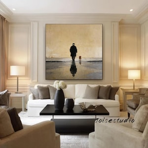 May include: A large framed painting depicts a person walking a dog on a wet surface, with a reflection of the figures. The artwork features a muted color palette of beige, gray, and black, creating a serene and contemplative atmosphere. The painting is displayed above a white sofa.