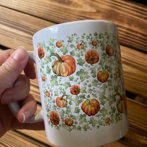 Peut inclure: Mug en céramique blanche orné de citrouilles oranges et de vignes vertes. La tasse a une anse incurvée et est conçue avec un motif répété de citrouilles de différentes tailles. C'est un article sur le thème de l'automne.
