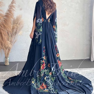 May include: A long, flowing navy blue dress with floral embroidery in red, yellow, and green. The dress features a low back and long, flowing sleeves. A black hat is worn.
