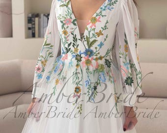 May include: White wedding dress with long sleeves and a V-neckline. The dress features colorful floral embroidery across the bodice and sleeves. The skirt is made of a flowing, sheer fabric. The text "Amber Bride" is visible at the bottom of the dress.