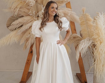 May include: A floor-length white gown featuring short puff sleeves, a round neckline, and a full, pleated skirt. The dress is made of a smooth, solid fabric and includes a train that extends along the floor. The text AmberBride is visible.