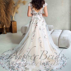 May include: A white wedding dress with floral embroidery. The dress features a V-neckline, cap sleeves, and a long train. The embroidery includes pink and green flowers. The text "Amber Bride" is visible on the dress.