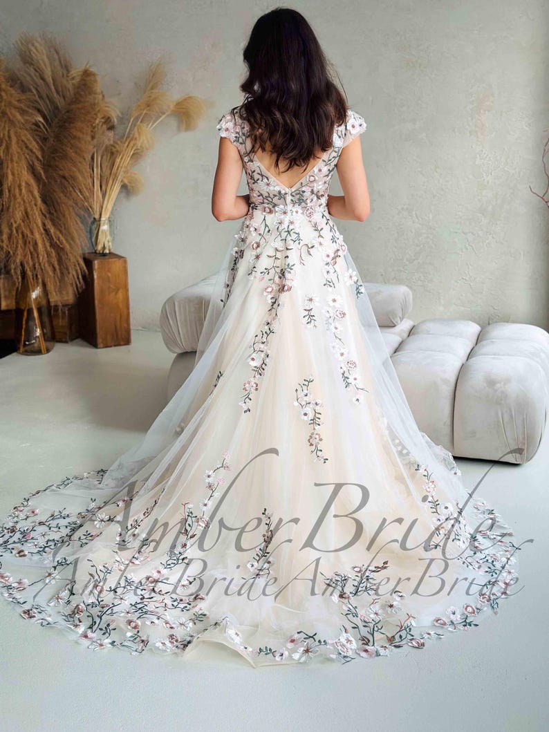 May include: A white wedding dress with floral embroidery. The dress features a V-neckline, cap sleeves, and a long train. The embroidery includes pink and green flowers. The text "Amber Bride" is visible on the dress.