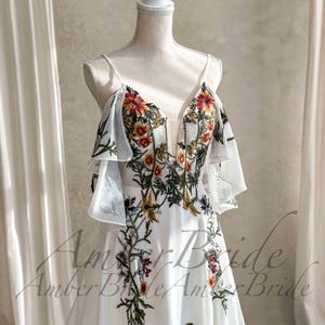 May include: A white wedding dress with floral embroidery in red, orange, yellow, and green. The dress features a deep V-neckline, spaghetti straps, and sheer, ruffled sleeves. The floral design cascades down the bodice and skirt.