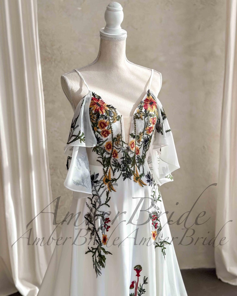 May include: A white wedding dress with floral embroidery in red, orange, yellow, and green. The dress features a deep V-neckline, spaghetti straps, and sheer, ruffled sleeves. The floral design cascades down the bodice and skirt.