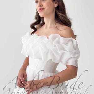May include: A white off-the-shoulder wedding dress with a ruffled neckline and short sleeves. The dress has a fitted bodice and a flowing skirt. The text "Amber Bride" is visible in the image.