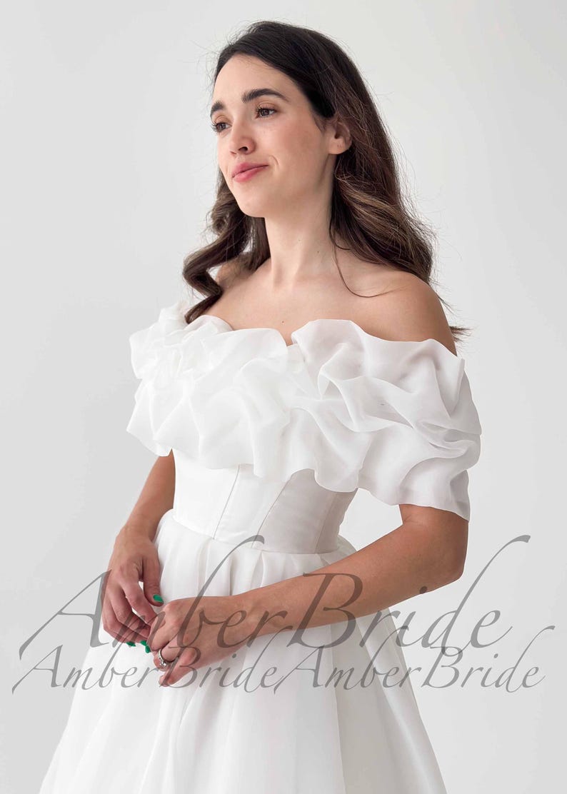 May include: A white off-the-shoulder wedding dress with a ruffled neckline and short sleeves. The dress has a fitted bodice and a flowing skirt. The text "Amber Bride" is visible in the image.