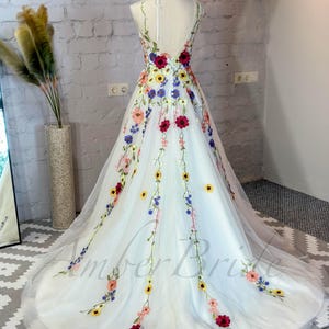 May include: A white tulle wedding dress with a floral embroidery design. The dress features a variety of colorful flowers, including red, yellow, blue, and pink. The dress has a V-neckline and a sleeveless design.