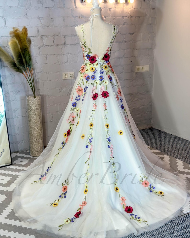 May include: A white tulle wedding dress with a floral embroidery design. The dress features a variety of colorful flowers, including red, yellow, blue, and pink. The dress has a V-neckline and a sleeveless design.