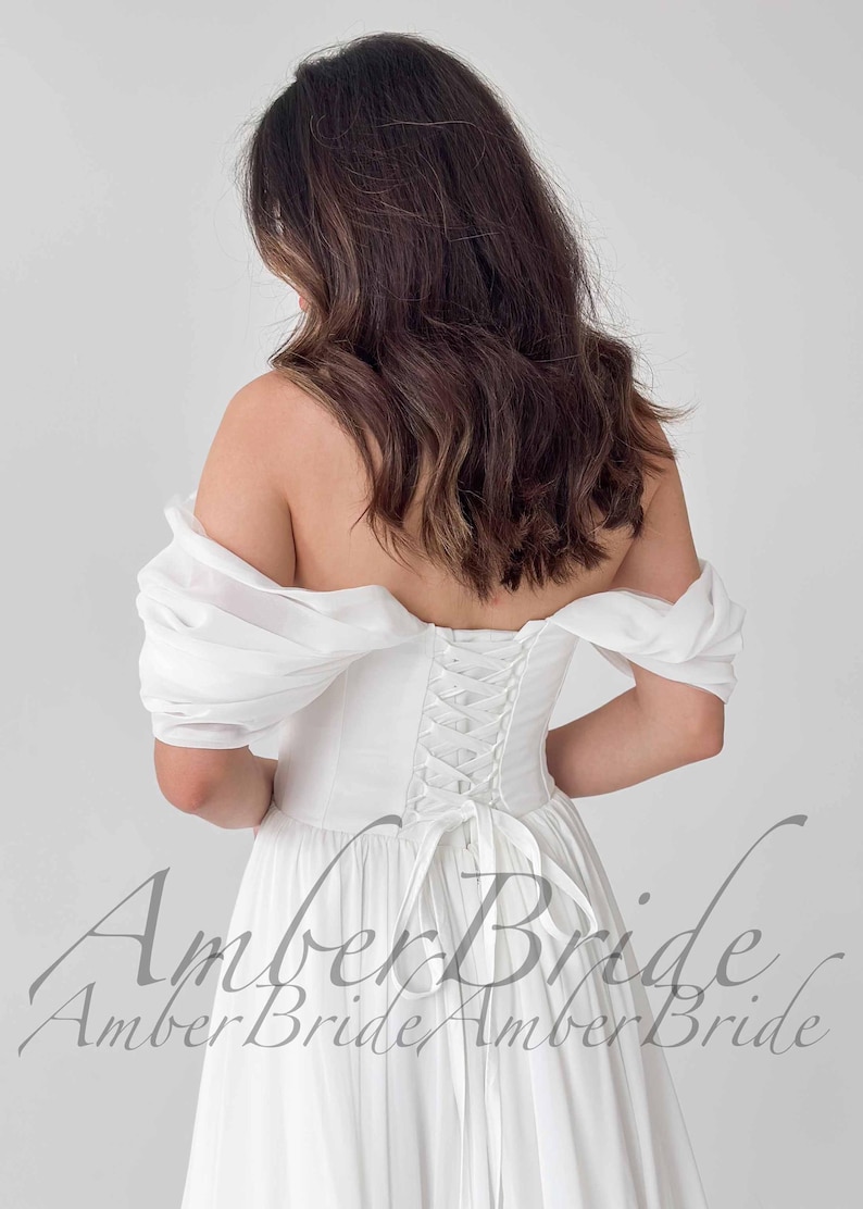 May include: A white off-the-shoulder wedding dress with a corset back and a flowing skirt. The dress features a lace-up back detail and short, puffy sleeves. The image shows the back of the dress, highlighting the design and fit.