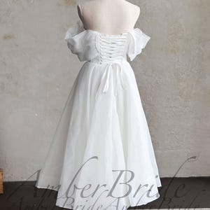 May include: A white, off-the-shoulder wedding dress on a mannequin. The dress features a lace-up back, short sleeves, and a knee-length skirt. The dress is displayed on a white mannequin with a wooden base. The background is a neutral color.