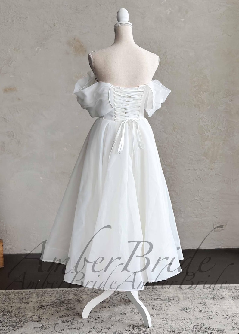 May include: A white, off-the-shoulder wedding dress on a mannequin. The dress features a lace-up back, short sleeves, and a knee-length skirt. The dress is displayed on a white mannequin with a wooden base. The background is a neutral color.