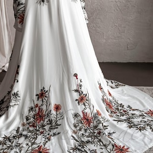 May include: A white maxi dress with long sleeves and a floral embroidery design. The embroidery features red, orange, and green flowers and leaves.