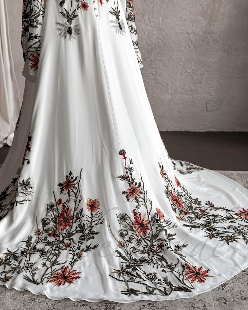 May include: A white maxi dress with long sleeves and a floral embroidery design. The embroidery features red, orange, and green flowers and leaves.