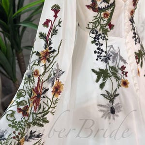 May include: White maxi dress with long sleeves and floral embroidery. The embroidery features a variety of flowers in shades of red, yellow, green, and black. The dress has a V-neckline and a button-up back.