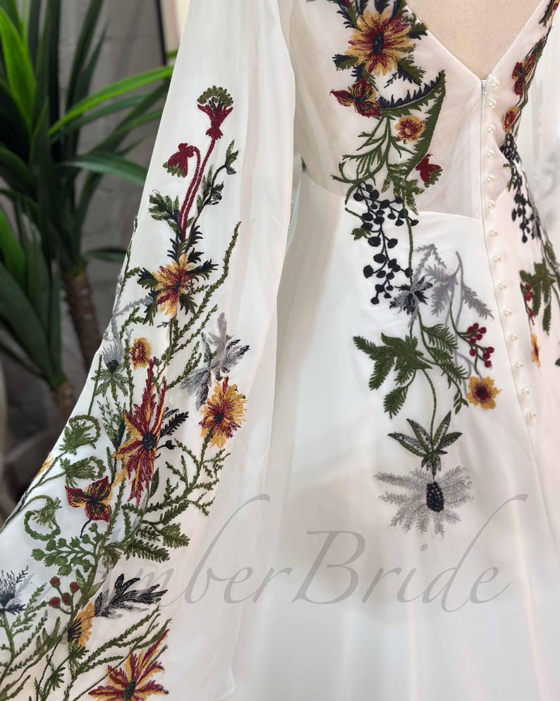 May include: White maxi dress with long sleeves and floral embroidery. The embroidery features a variety of flowers in shades of red, yellow, green, and black. The dress has a V-neckline and a button-up back.