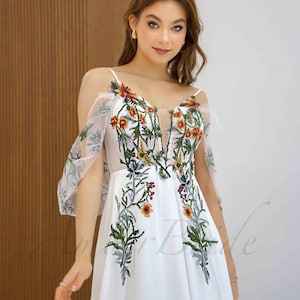 May include: White maxi dress with a floral embroidery design. The dress has a V-neckline, thin straps, and sheer, ruffled sleeves.