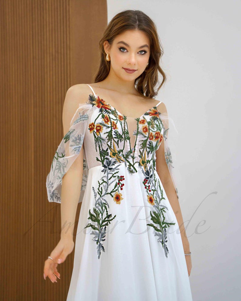 May include: White maxi dress with a floral embroidery design. The dress has a V-neckline, thin straps, and sheer, ruffled sleeves.