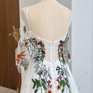 May include: White wedding dress with a floral embroidery design. The dress has a sweetheart neckline and a fitted bodice. The skirt is full and flowing. The back of the dress features a row of pearl buttons.
