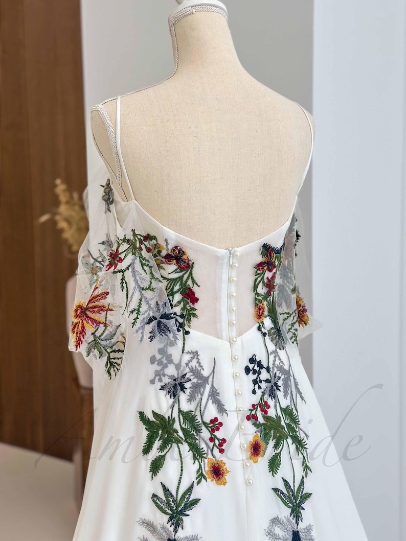 May include: White wedding dress with a floral embroidery design. The dress has a sweetheart neckline and a fitted bodice. The skirt is full and flowing. The back of the dress features a row of pearl buttons.