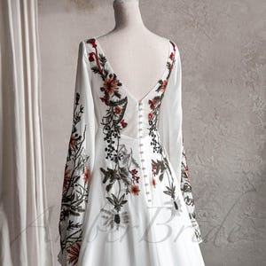 May include: A white wedding dress with long sleeves and a V-neckline. The dress is adorned with floral embroidery in shades of red, green, and brown.
