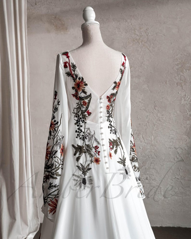 May include: A white wedding dress with long sleeves and a V-neckline. The dress is adorned with floral embroidery in shades of red, green, and brown.
