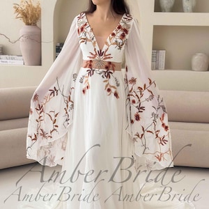 May include: A white wedding dress with a plunging neckline and long, sheer sleeves. The dress features floral embroidery in shades of brown and red. The text "Amber Bride" is visible at the bottom of the dress.