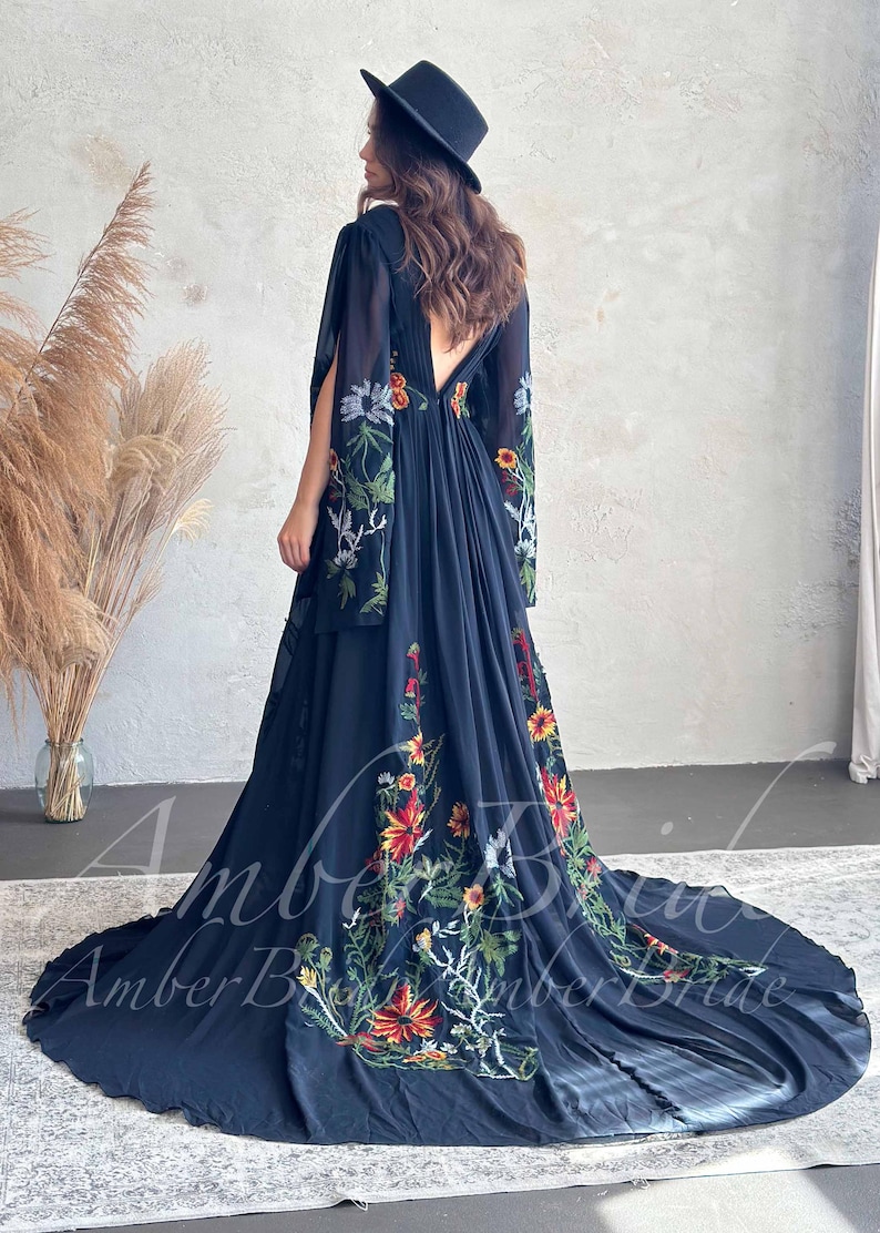 May include: A long, flowing navy blue dress with floral embroidery in red, yellow, and green. The dress features a low back and long, flowing sleeves. A black hat is worn.