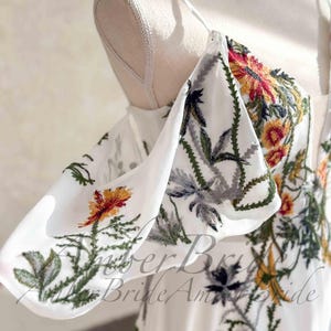 May include: White off-the-shoulder dress with floral embroidery in red, yellow, and green. The dress features a square neckline and spaghetti straps. The embroidery includes flowers and leaves. The text "Amber Bride Am" is visible.