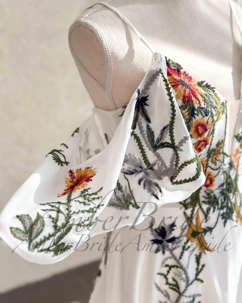 May include: White off-the-shoulder dress with floral embroidery in red, yellow, and green. The dress features a square neckline and spaghetti straps. The embroidery includes flowers and leaves. The text "Amber Bride Am" is visible.