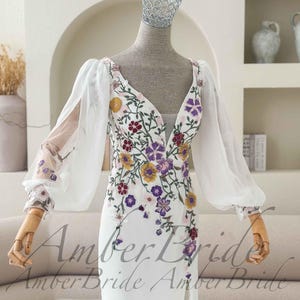 May include: White wedding dress with a plunging neckline and floral embroidery in yellow, purple, and burgundy. The dress features sheer, long sleeves and a high slit. The text "Amber Bride" is visible at the bottom of the dress.