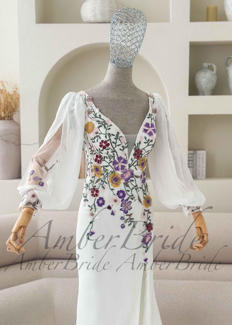 May include: White wedding dress with a plunging neckline and floral embroidery in yellow, purple, and burgundy. The dress features sheer, long sleeves and a high slit. The text "Amber Bride" is visible at the bottom of the dress.