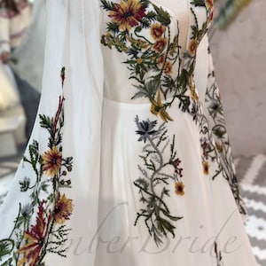 May include: White maxi dress with long sleeves and a V-neckline. The dress is adorned with colorful floral embroidery.