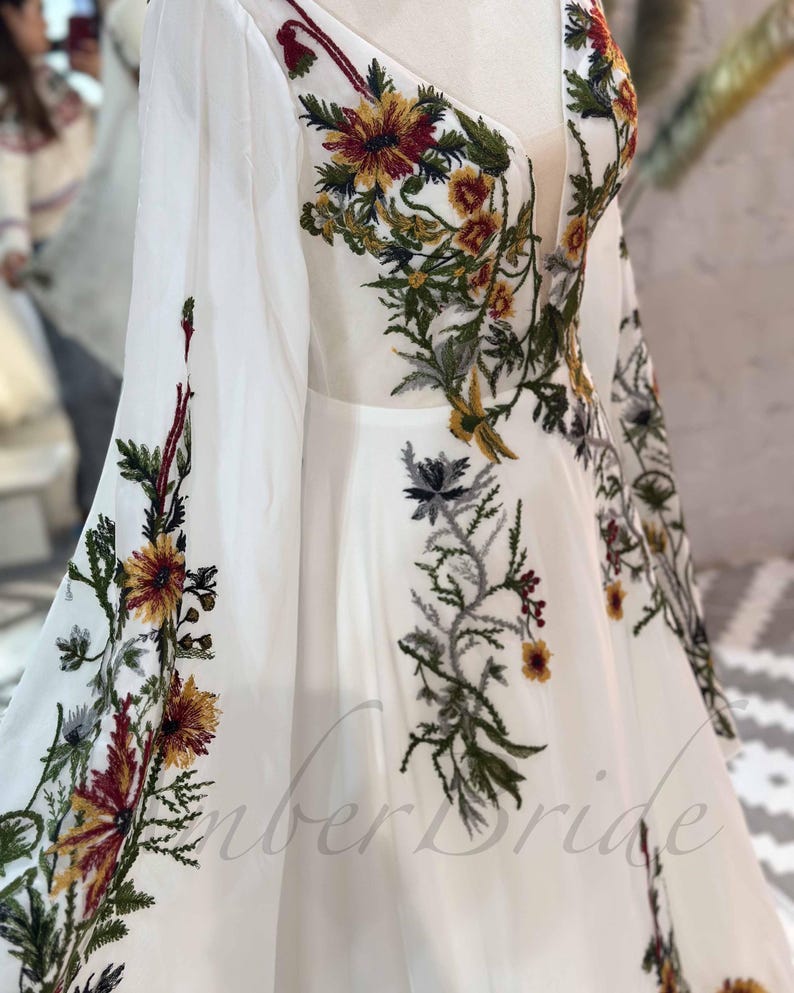 May include: White maxi dress with long sleeves and a V-neckline. The dress is adorned with colorful floral embroidery.