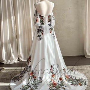 May include: A white wedding dress with floral embroidery. The dress features off-the-shoulder sleeves and a flowing skirt. The floral design includes red, orange, and green accents. The dress is displayed on a mannequin.
