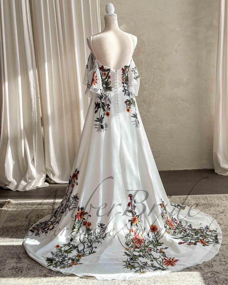 May include: A white wedding dress with floral embroidery. The dress features off-the-shoulder sleeves and a flowing skirt. The floral design includes red, orange, and green accents. The dress is displayed on a mannequin.