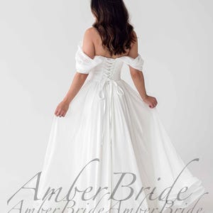 May include: A white off-the-shoulder wedding dress with a corset back and flowing skirt. The dress features a lace-up back and a full, floor-length skirt. The image shows the back of the dress, with the text "Amber Bride" in a script font.