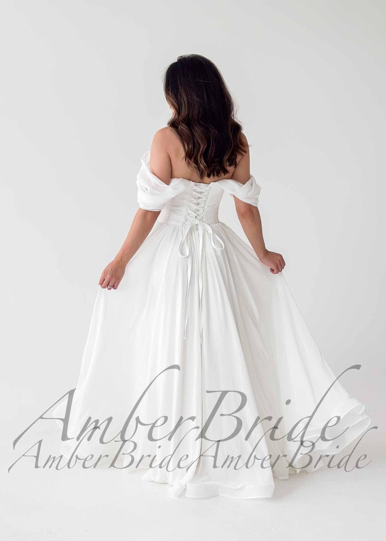 May include: A white off-the-shoulder wedding dress with a corset back and flowing skirt. The dress features a lace-up back and a full, floor-length skirt. The image shows the back of the dress, with the text "Amber Bride" in a script font.