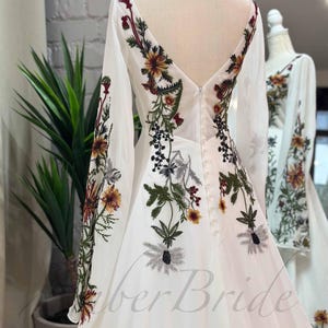May include: White wedding dress with long sleeves and a V-neckline. The dress is adorned with floral embroidery in shades of green, red, and yellow.