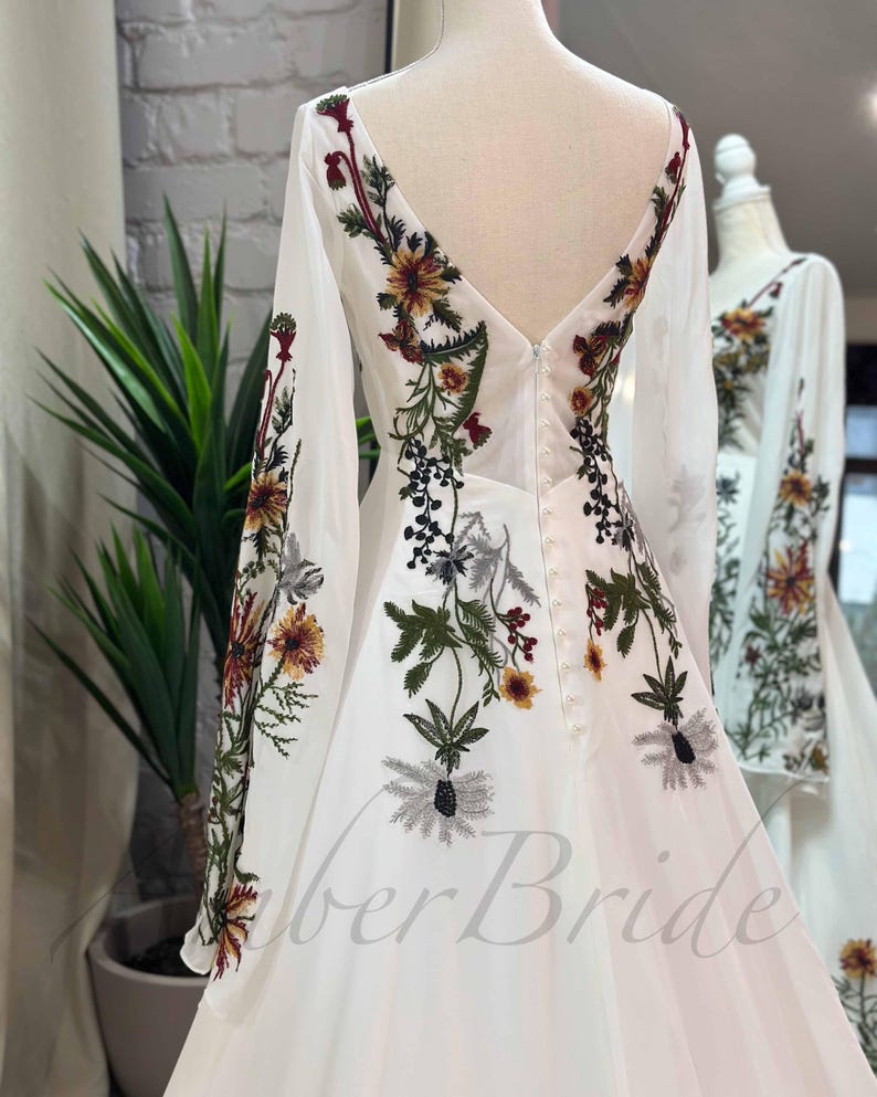 May include: White wedding dress with long sleeves and a V-neckline. The dress is adorned with floral embroidery in shades of green, red, and yellow.