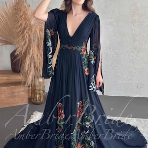 May include: A black, floor-length dress with a plunging neckline and long, flowing sleeves. The dress features colorful floral embroidery along the sleeves, bodice, and skirt. A woman is wearing a black hat and holding her hand up.