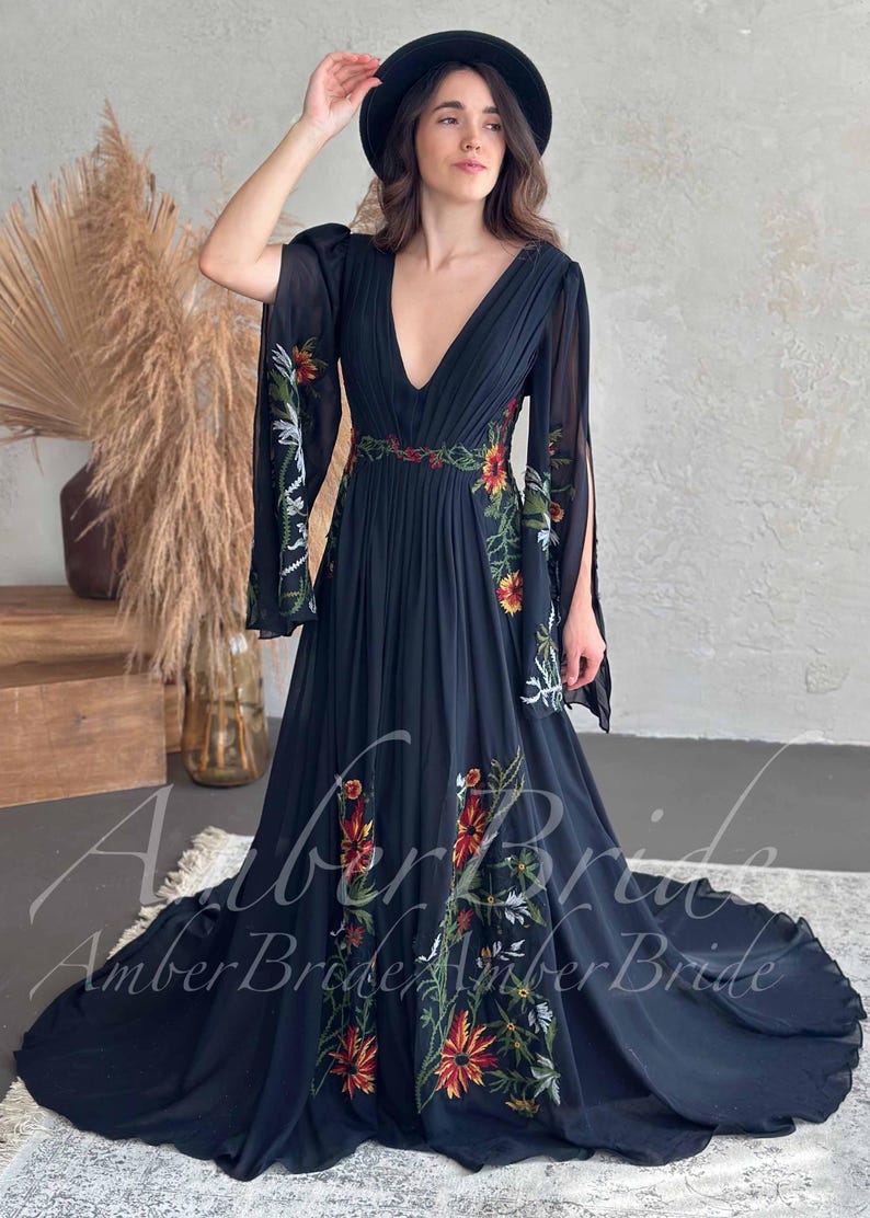 May include: A black, floor-length dress with a plunging neckline and long, flowing sleeves. The dress features colorful floral embroidery along the sleeves, bodice, and skirt. A woman is wearing a black hat and holding her hand up.
