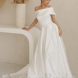May include: A white off-the-shoulder wedding dress with a flowing skirt and a beaded waistband. The dress is photographed on a model in a studio setting.
