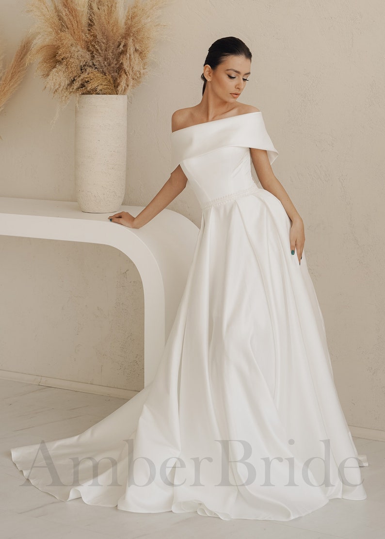 May include: A white off-the-shoulder wedding dress with a flowing skirt and a beaded waistband. The dress is photographed on a model in a studio setting.