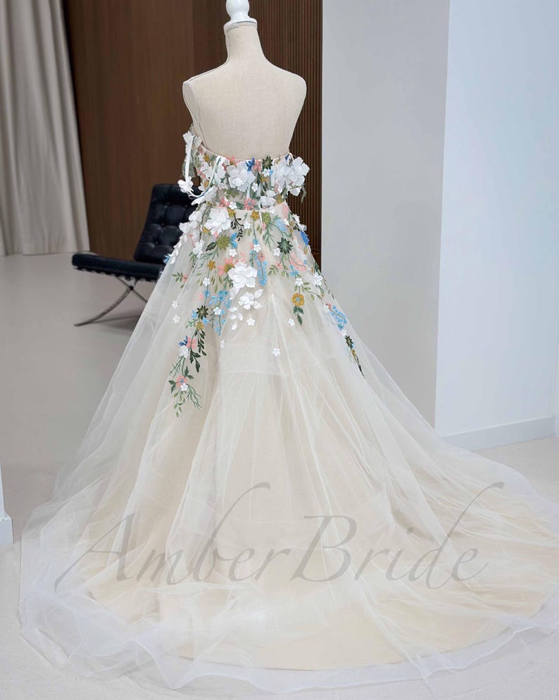 May include: A white tulle wedding dress with a strapless bodice and a floral embroidery design. The dress has a full skirt and a long train.