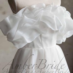 May include: A white off-the-shoulder wedding dress with a layered, ruffled neckline. The bodice is fitted, and the skirt flows down. The dress is displayed on a mannequin. The text "Amber Bride" is visible at the bottom.