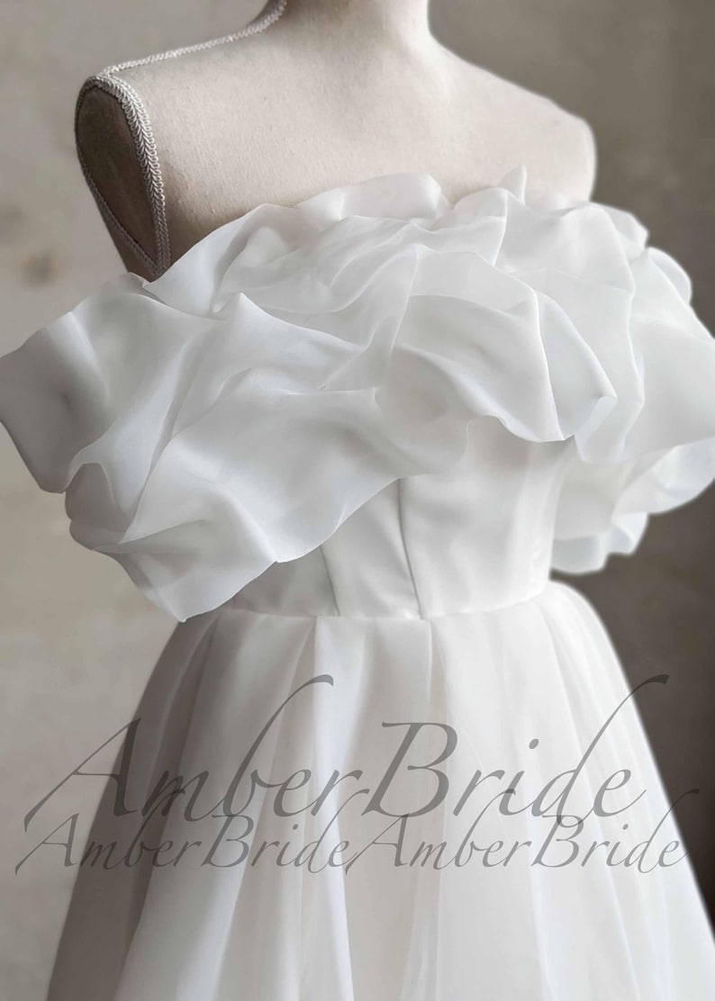 May include: A white off-the-shoulder wedding dress with a layered, ruffled neckline. The bodice is fitted, and the skirt flows down. The dress is displayed on a mannequin. The text "Amber Bride" is visible at the bottom.